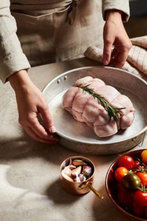 A butcher prepares raw meat adorned with fresh rosemary in a metal container. The scene is