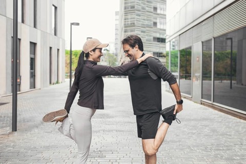 A couple dressed in sportswear performs a leg stretch on a city street, supporting each other with