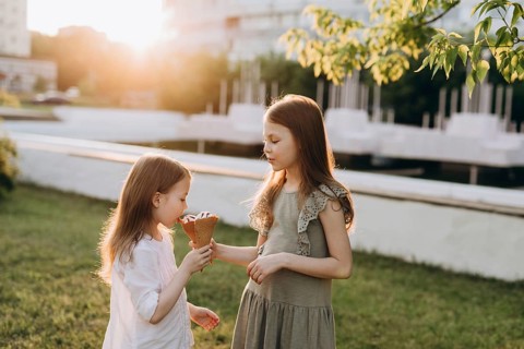 Two young girls in a sunlit park share an ice cream cone, enjoying a warm summer day. The scene
