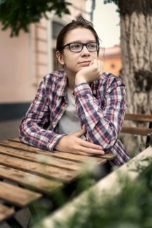 A teenage boy with long hair and glasses wearing a plaid shirt sits thoughtfully at an outdoor
