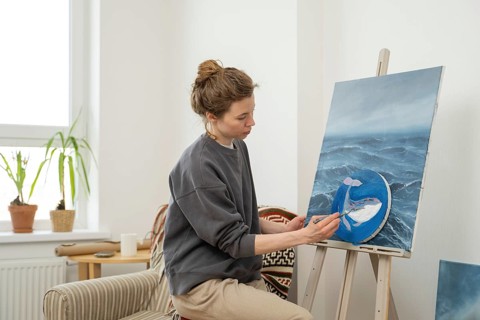 An artist in a gray sweatshirt creates a vibrant ocean themed painting on an easel. The cozy room