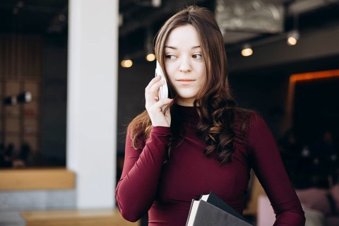 A young female student stands indoors, balancing books while talking on her phone. Her expression
