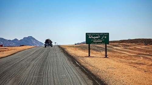 An off-road vehicle on a sandy road next to a sign in a vast desert environment, By car through the