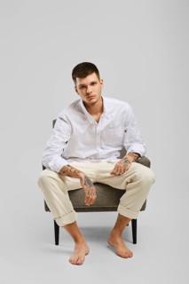 Full length studio portrait of young barefoot man in white shirt and ivory trousers sitting in