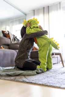 Mother kneeling on carpet and tenderly hugging her son wearing a dinosaur costume, in a cozy home