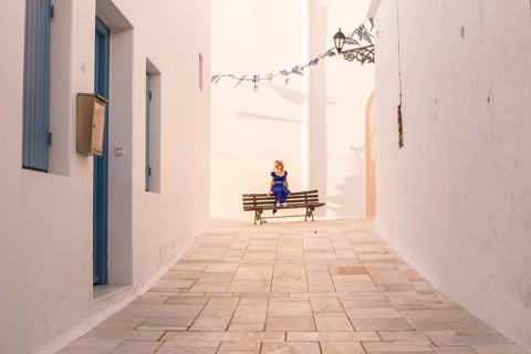 A woman in a blue dress sits on a bench in a sunlit alley of a Greek village in Plaka, Milos island