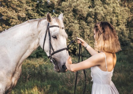 A young woman gently holds the reins of a white horse in a sunlit meadow. Her white dress