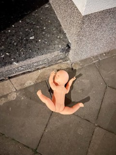 An undressed doll lies on the pavement in front of a house, symbolic image