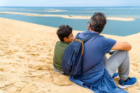 A father and son sit on the dune of Pilat, overlooking the serene ocean. The moment captures
