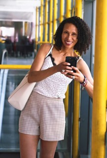 A Latin woman stands in a bright corridor, smiling and engaged with her cell phone. She wears