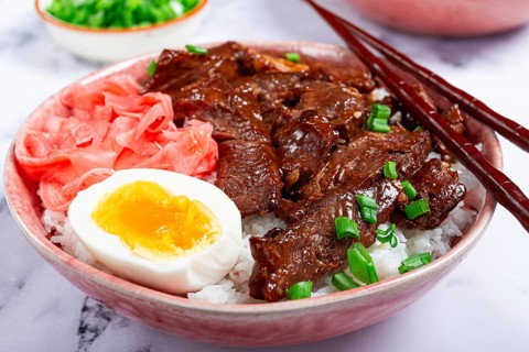 Gyudon Japanese beef with rice, pickled ginger, boiled egg slice, garnished with green onions, in a