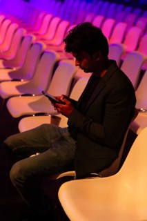 A indian man in a dark blazer and jeans sits in an empty, illuminated auditorium at a conference of
