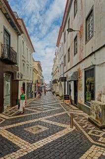 People walk along a narrow cobbled street lined with historic buildings and cafés under a clear