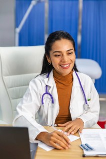 A female doctor with a stethoscope around her neck sits at a desk, writing notes and smiling. The