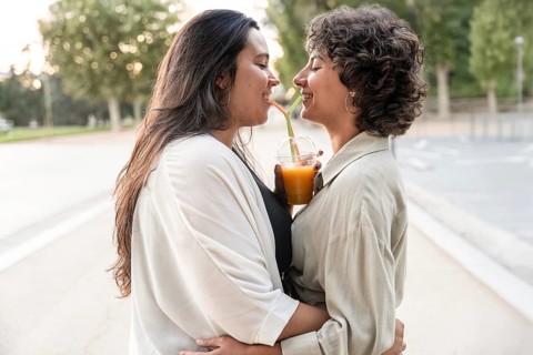 A lesbian couple embraces and shares a drink outdoors. Smiling, they enjoy each other's company in