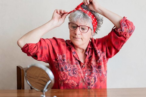 A elderly woman in a vibrant red, floral shirt adjusts her red polka dot headscarf while looking in