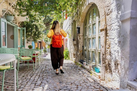A female tourist with a red backpack strolls through a charming, sunlit cobblestone street. Lush