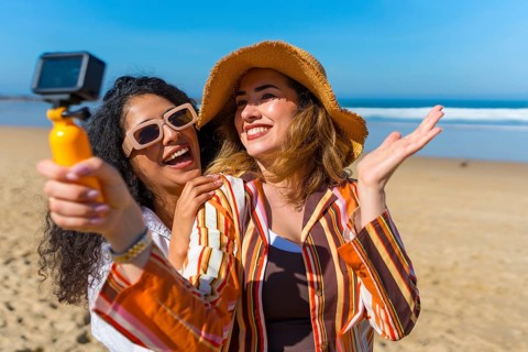 Two smiling young women creating content with a small video camera on a sunny sandy beach, having