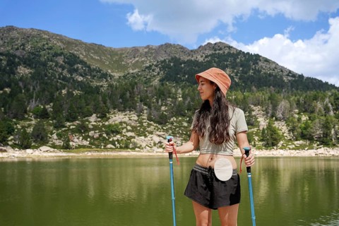 A woman with an ostomy bag stands confidently by a serene mountain lake, wearing a hat and holding