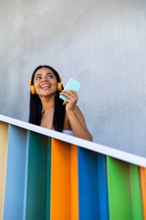 A joyful woman with headphones smiles while using her smartphone against a backdrop of vibrant