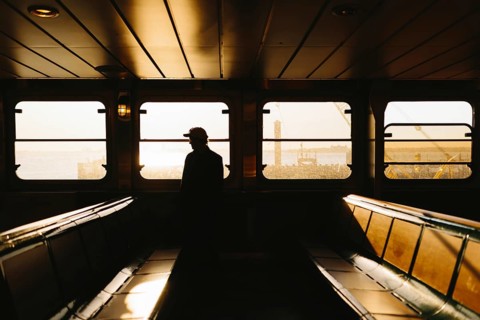A man in silhouette stands inside a ferry, gazing out at a serene, sunlit harbor scene in New York.