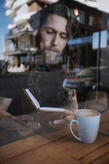 A man with long hair reads a book while enjoying a hot drink at a cafe. The view through the window