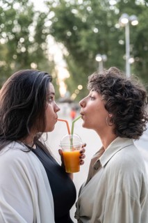 A lesbian couple shares a juice through straws in a tender moment, standing outdoors with a