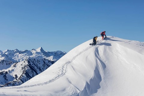 Two snowboarder friends make their way up a pristine snow covered peak, leaving tracks behind. The