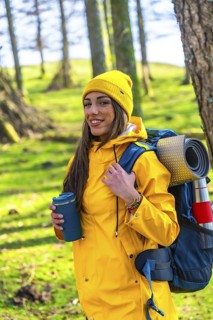 Young hiker woman wearing a yellow raincoat and backpack is holding a hot drink in a forest during