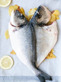 Two raw whole sea bream are shaped like a heart, ready for baking with potatoes and lemon