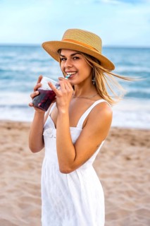 Young woman wearing straw hat and white dress drinking refreshing beverage on beach during summer