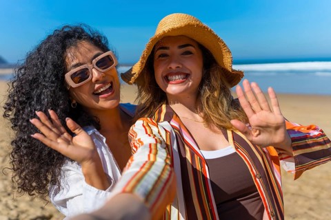 Happy women friends enjoying summer beach vacation, smiling and waving at the camera while taking a