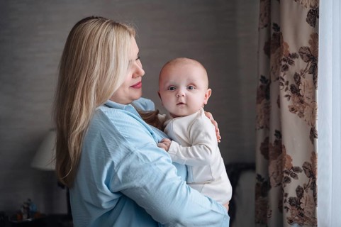A mother in a light blue outfit cradles her baby boy by a window. The room has a cozy ambiance with