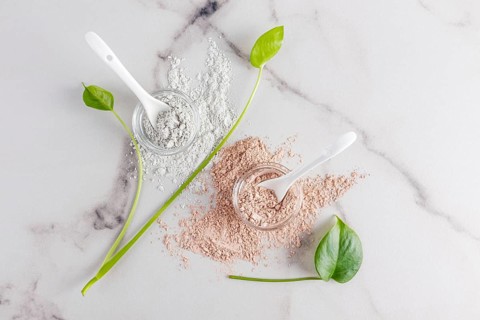 Top view of natural cosmetic clay powders in clear bowls with spoons on a marble surface,