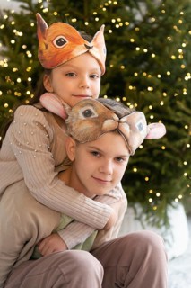 A girl and boy embrace, wearing animal masks, with a beautifully lit Christmas tree in the