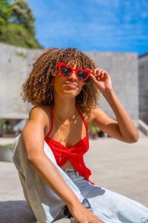 Vertical photo of a cute latin woman with curly hair using heart shape red sunglasses sitting in