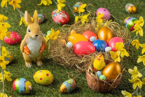 A rabbit figure stands next to a nest full of colorful Easter eggs, decorated with yellow flowers