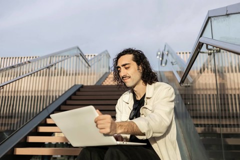 A young man with curly hair sits on stairs outdoors, working on a laptop Sunlight casts a serene