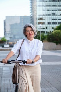 A mature woman with gray hair stands confidently with a scooter in an urban environment. She wears