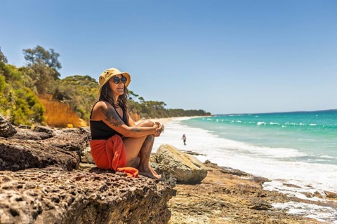 A woman sits on rocks, enjoying the serene beach view under a clear blue sky in Jervis Bay,