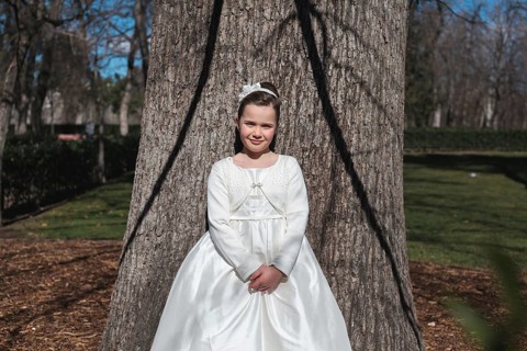 A young girl wearing a beautiful First Communion dress stands by a large tree. The serene outdoor