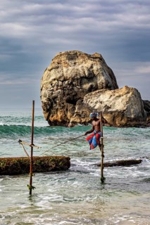 A fisherman sits on a wooden pole in the water, in front of a majestic rocky landscape, The stilt