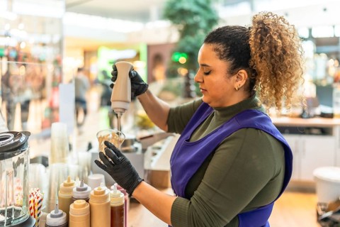Barista in apron and gloves carefully squeezes sauce into a cup, crafting a sweet beverage at a