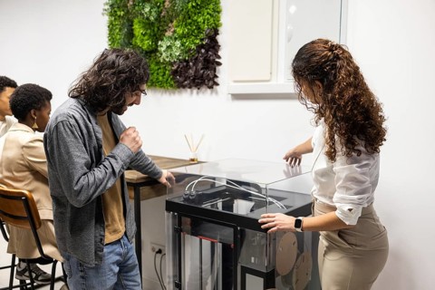 A diverse team collaborates in a modern office. Colleagues engage with a 3D printer while others