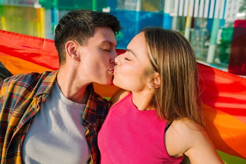 A passionate lesbian couple shares a romantic kiss beneath a flowing rainbow Pride flag. Their