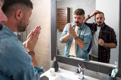 A gay couple stands together in front of a bathroom mirror, engaging in their morning routine. They