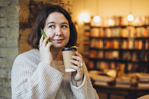 A woman in a relaxed sweater holds a coffee cup and green smartphone while sitting in a bookstore