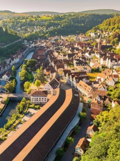 Aerial view of a town with many roofs and a river and bridge, surrounded by green hills and