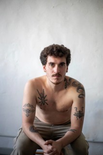 A young man with a short beard and curly hair sits in a relaxed pose against a plain white wall,