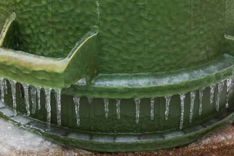 Upside down green PVC and rubber thrash can covered with thick sheet of ice and hanging icicles